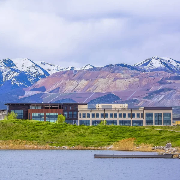 Square Buildings and snowy mountain beyond the grassy shore of a lake on a cloudy day