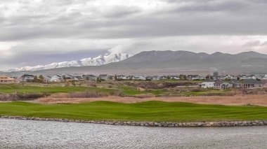 Çimenli kıyının ötesinde evler ve karla kaplı dağ ile Panorama Manzara göl manzarası
