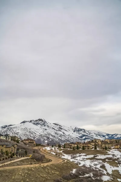 Kıvrımlı yol ve yükselen kar kaplı dağ manzaralı bir tepe üzerine inşa edilmiş evler — Stok fotoğraf