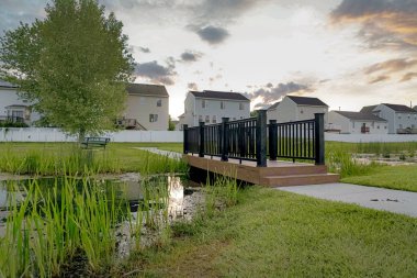 Gün batımında bir ağaç evler ve coudy gökyüzü manzaralı küçük bir gölet üzerinde Köprü