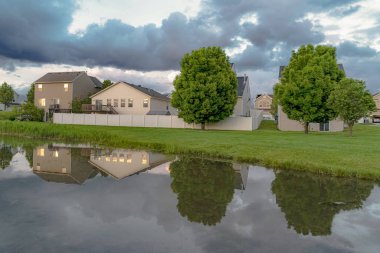 Evler ve ağaçlar çimenli bir arazi ortasında bir göletin parlak su yansıyan