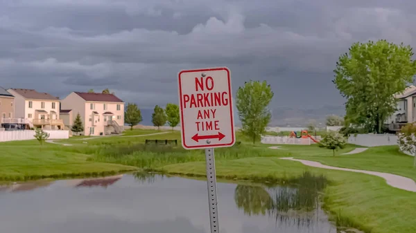 Marco panorámico No Parking En Cualquier Momento signo con estanque terreno herboso y casas en el fondo — Foto de Stock