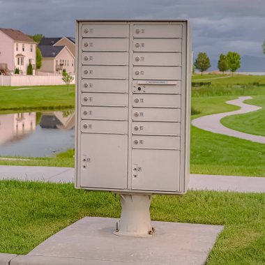 Square frame Cluster mailbox against pond and houses under sky with thick gray clouds clipart