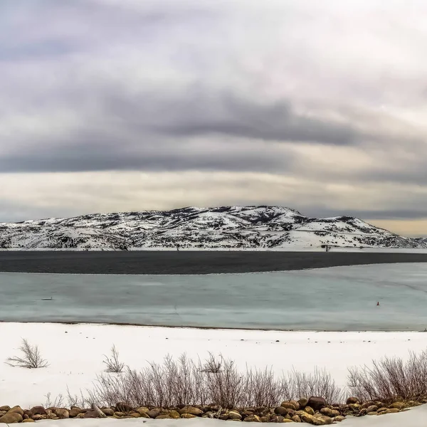 Čtvercový rám panoramatický výhled na zamrzlé jezero a zasněžené hory v zimě pod zamračeným nebem — Stock fotografie