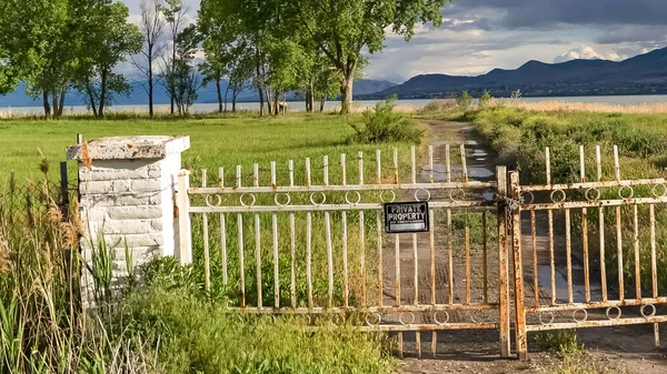 Panorama Rusty gate chain link fence and dirt road on a grassy field in front of a lake