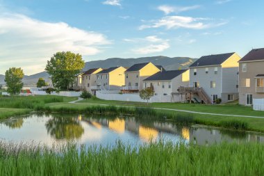 Bulutlu gökyüzü altında parlak gölet ve yükselen dağ manzaralı evler