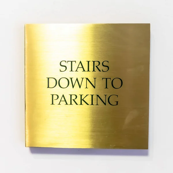 Square Gold plated sign that reads Stairs Down To Parking against white interior wall
