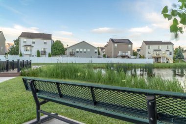 Bulutlu gökyüzü altında çimenli gölet ve evler manzaralı Yeşil metal park tezgah