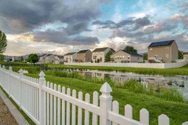 Gölet ve evler beyaz ahşap bir çitin diğer tarafında, gökyüzü bulutlu.