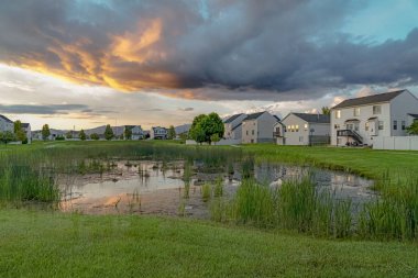 Bulutlu gökyüzünün altında çimenlerin ve evlerin ortasındaki göletin manzaralı gün batımı manzarası