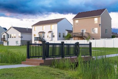 Gökyüzü bulutlu evlerin önündeki çimenli bir parkta köprü ve gölet.