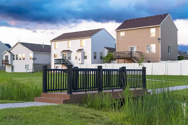 Gökyüzü bulutlu evlerin önündeki çimenli bir parkta köprü ve gölet.