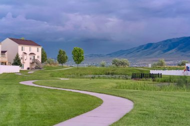 Bulutlu gökyüzünün altında gölü ve köprüsü olan çimenli bir parkta esen yol.