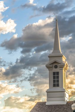 Mavi gökyüzüne ve gri kabarık bulutlara karşı bir kilisenin beyaz çan kulesi ve karanlık çatısı