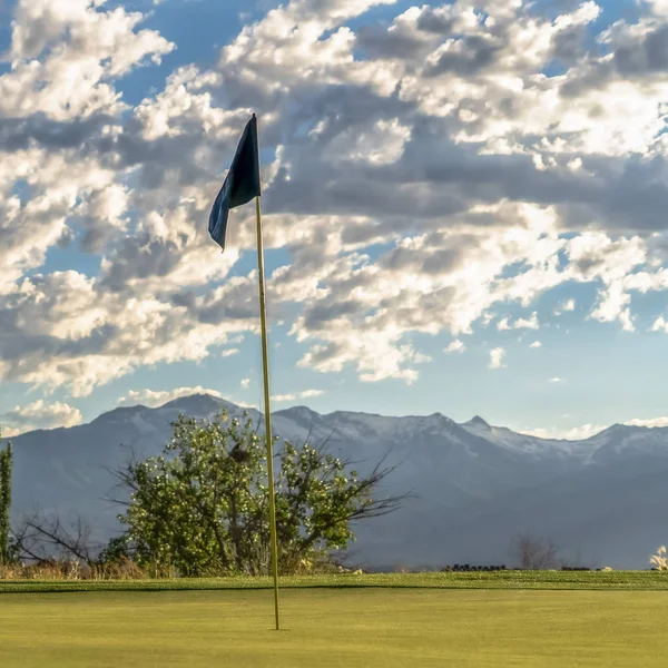 Marco cuadrado Enfoque en el asta de la bandera de un campo de golf contra árboles montaña y cielo nublado — Foto de Stock