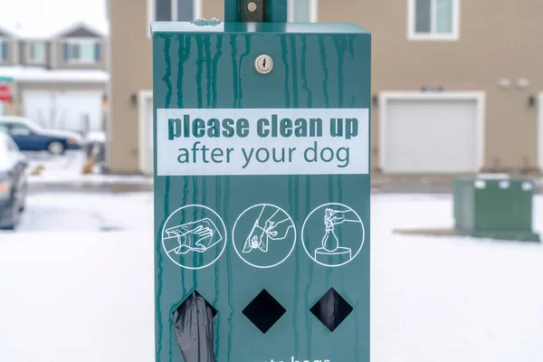 Station de déchets pour animaux de compagnie avec sacs de crotte de chien noir contre les maisons floues et le sol enneigé — Photo