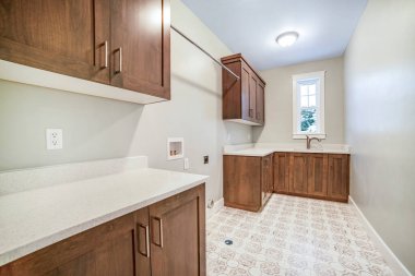 Kahverengi ahşap dolaplı mutfak. Beyaz tezgahlar lavabo ve pencere.