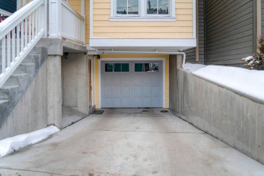 Gri camlı garaj kapısı ve verandaya çıkan merdivenleri olan karlı bir ev.