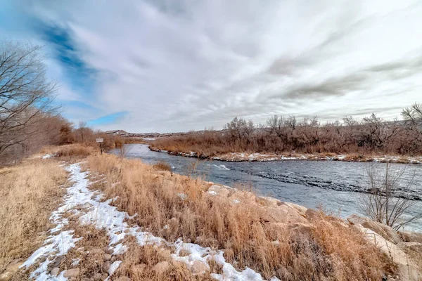 Flod med rinnande vatten längs gräsbevuxna och steniga stränder dammade av vintersnö — Stockfoto