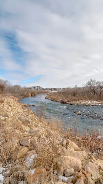 Vertical frame Scenic river with snowy shore against distant hills under cloudy sky in winter