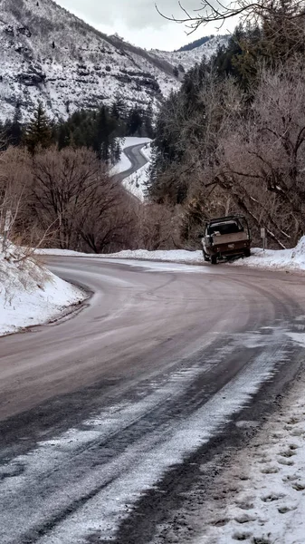 Vertical frame Road winding through snowy mountain with leafless trees and evergreens in winter