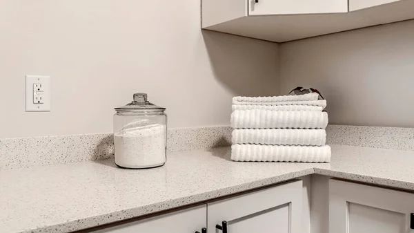 Panorama frame Countertop with jar of powder detergent and folded towels over wood cabinets