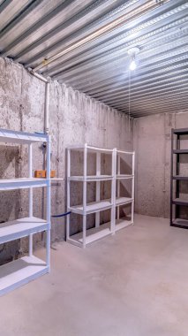 Beton duvar ve zemin olan bir depoda dikey olarak çok katmanlı metal raflar var.