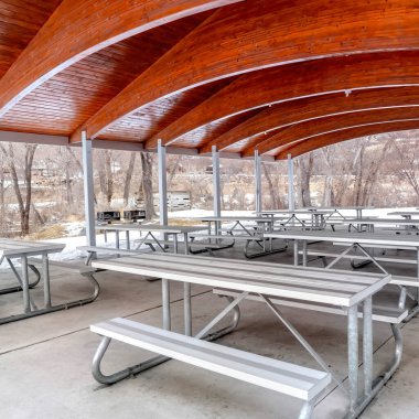 Tavanda ahşap kemerler olan bir park köşesinde kare masa ve banklar.