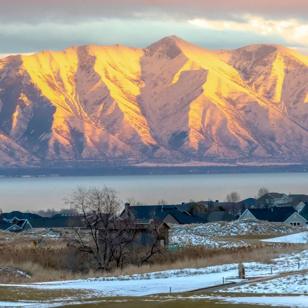 Cadre carré Montagne enneigée éclairée par le soleil doré au coucher du soleil avec lac et maisons au premier plan — Photo