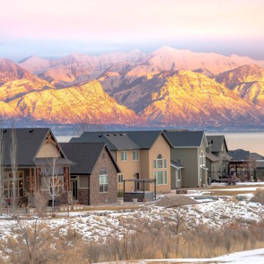 Karlı, güneşli dağları olan ve gün batımında sakin gölü olan evler cephesi.