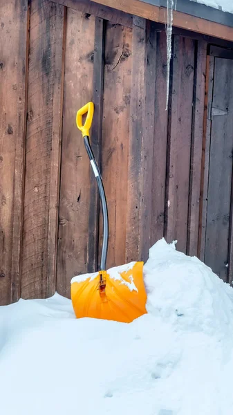 Vertical Yelllow snow shovel against snowed in ground and brown wooden wall of building — Stock Photo, Image