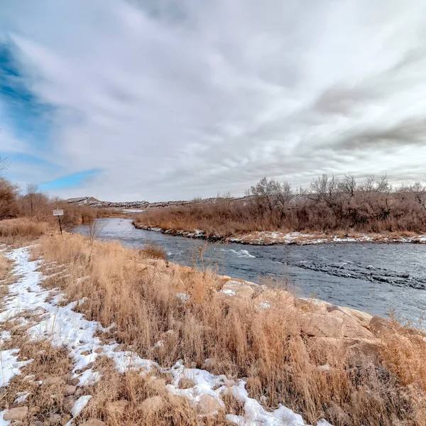 Río cuadrado con agua que fluye a lo largo de las orillas cubiertas de hierba y rocosas polvoreadas con nieve de invierno — Foto de Stock