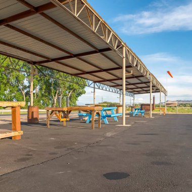 Bir parktaki köşeli bir çadırın düz kıvrımlı metal çatısının altında kare ekin masaları ve banklar