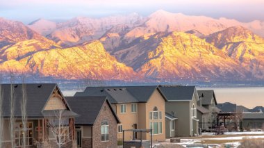 Panorama çerçeve karlı güneşli dağ manzaralı evler ve gün batımında sakin göl arka planı