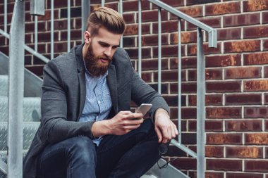 Yakışıklı sakallı adam açık havada elinde akıllı telefon ile merdivenlerde oturuyor. Şehirdeki şık adam.