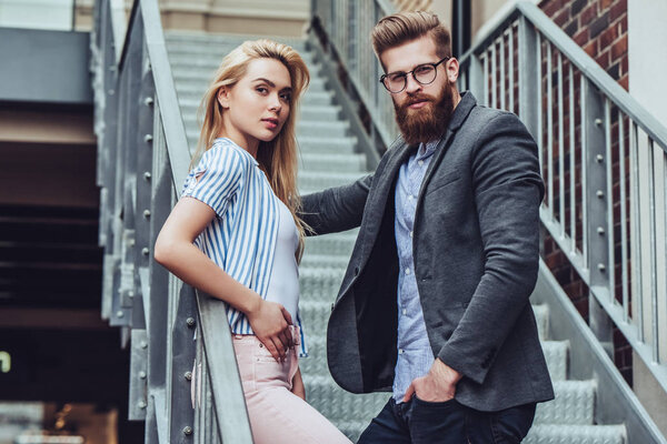 Attractive young happy couple is spending time together outdoors. Attractive woman and handsome bearded man are standing on stairs in the city.