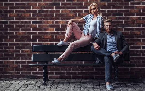 Stylish young couple is spending time together outdoors. Attractive woman and handsome bearded man are sitting on a bench on brick background in the city.