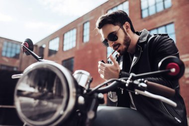 Yakışıklı sakallı motorcu klasik tarzı siyah motosiklet üzerinde otururken yasaktır sigarette var. Cafe racer.