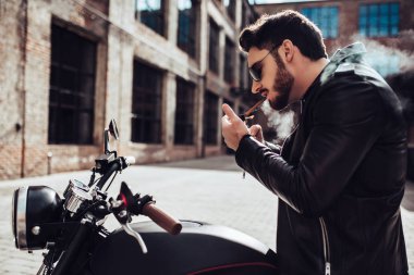 Yakışıklı sakallı motorcu klasik tarzı siyah motosiklet üzerinde otururken yasaktır sigarette var. Cafe racer.