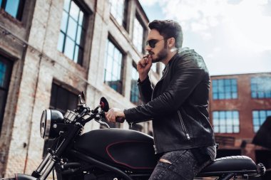 Yakışıklı sakallı motorcu klasik tarzı siyah motosiklet üzerinde otururken yasaktır sigarette var. Cafe racer.
