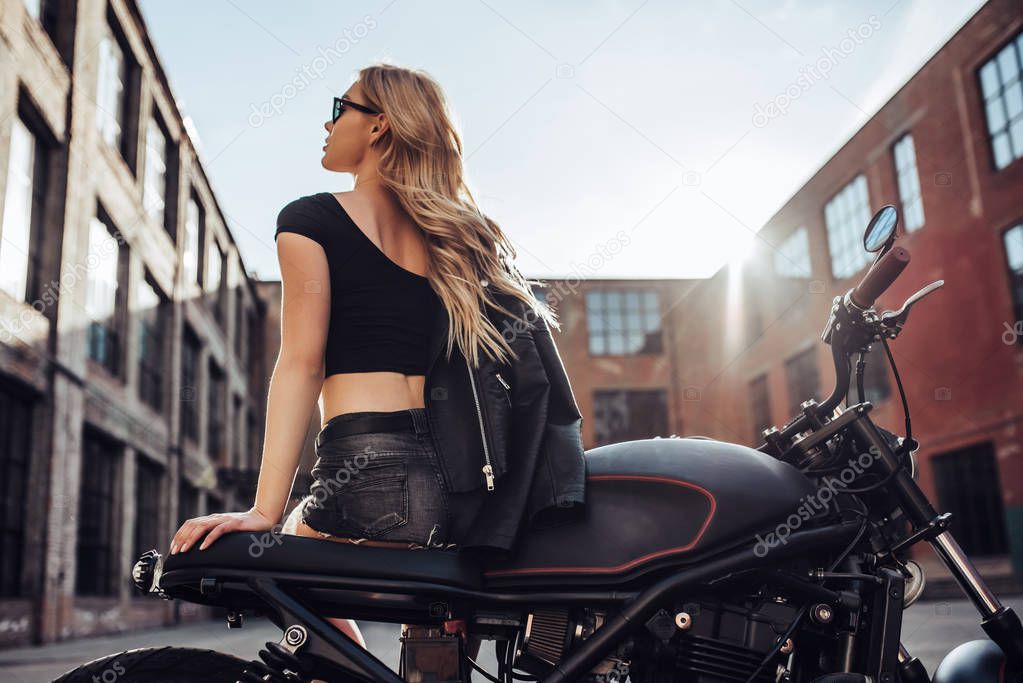 Attractive young woman in black leather outfit with classic style motorcycle. Female biker outdoors with cafe racer.