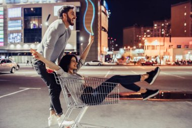 Genç romantik çift süpermarket arabası ile akşam eğleniyor. Sakallı yakışıklı ve çekici genç kadın birlikte dışarıda vakit.