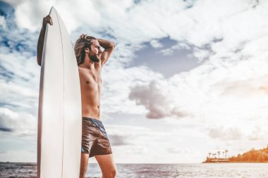 Uzun saçlı yakışıklı delikanlı elinde beyaz surfboard ile plajda durup.