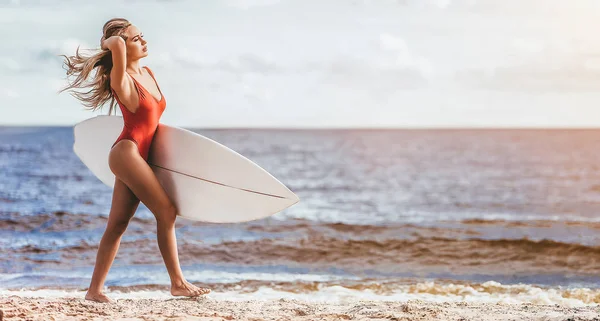 性感的年轻美女在红色泳装走在海滩 手中的白色冲浪板 — 图库照片