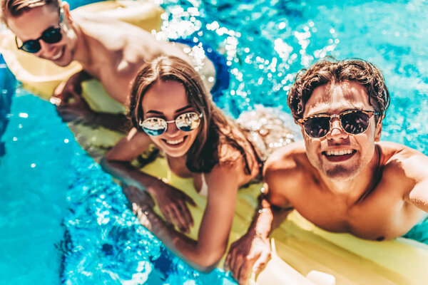 Happy attractive young friends in sunglasses are having fun in swimming pool on an inflatable mattress and smiling while making selfie.