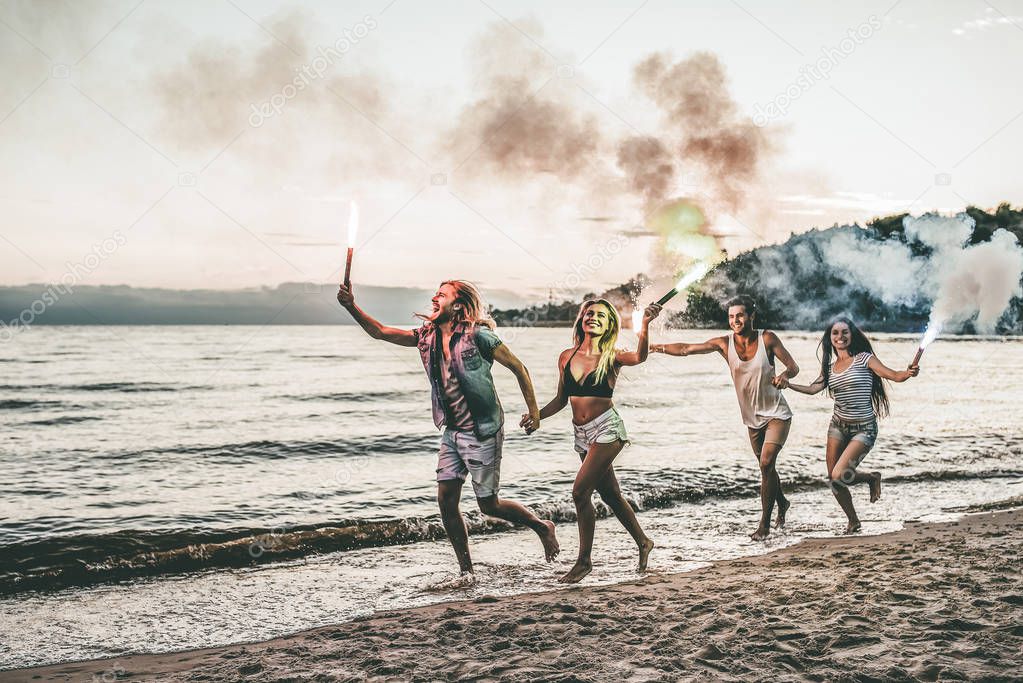 Group of young friends are having fun on beach with coloured smoke fires.