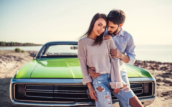 Romantic couple is standing near green retro car on the beach. Handsome bearded man and attractive young woman with vintage classic car. Love story.