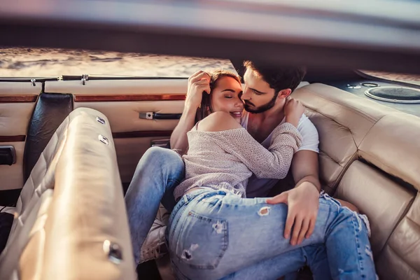 Romantic couple is sitting in green retro car on the beach. Handsome bearded man and attractive young woman in vintage classic car. Stylish love story. Hugging and kissing while being in car.