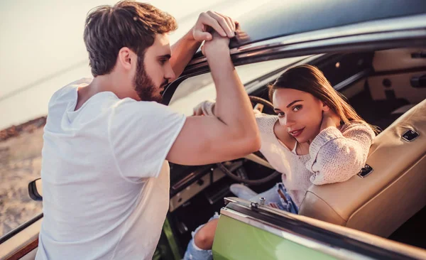 Romantic couple with green retro car on the beach. Handsome bearded man and attractive young woman with vintage classic car. Stylish girl is sitting in car while her man is standing near and is gonna kiss her. Love story.