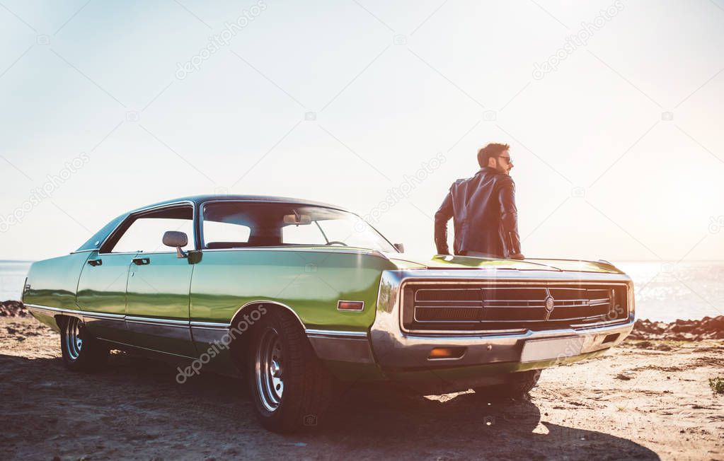 Handsome bearded man is standing near his green retro car on the beach. Vintage classic car.